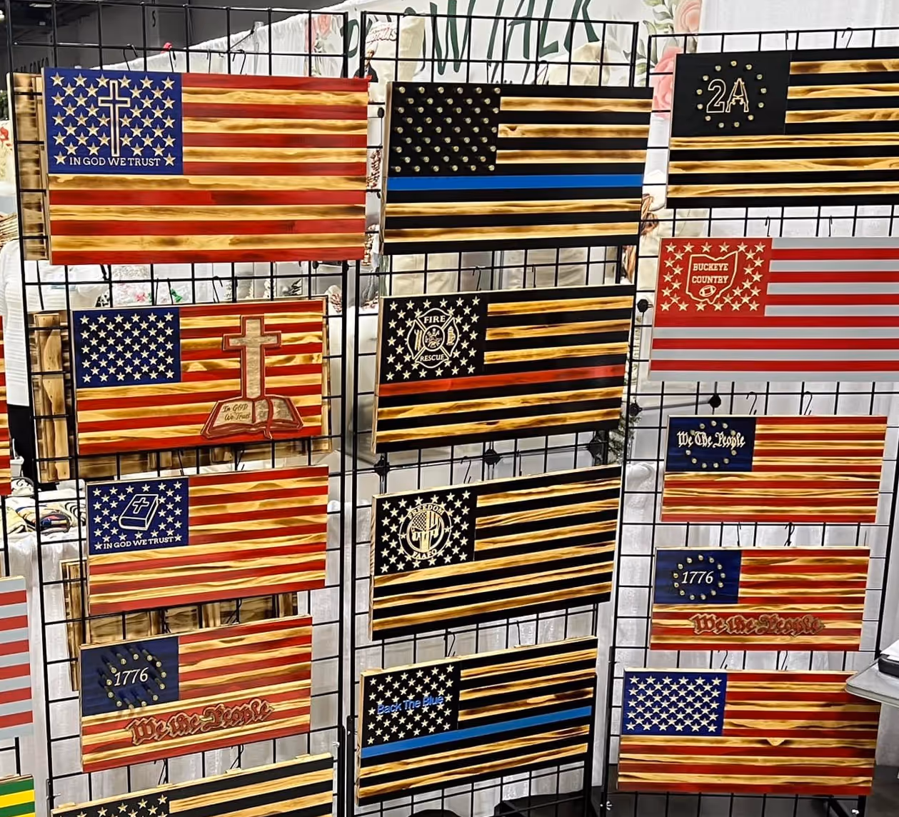 Various styled wooden American flags with religious, patriotic, and firefighter themes displayed on metal grids.