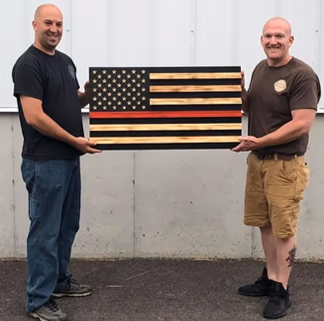 Two men holding a wooden American flag with a thin red line in front of a concrete wall.