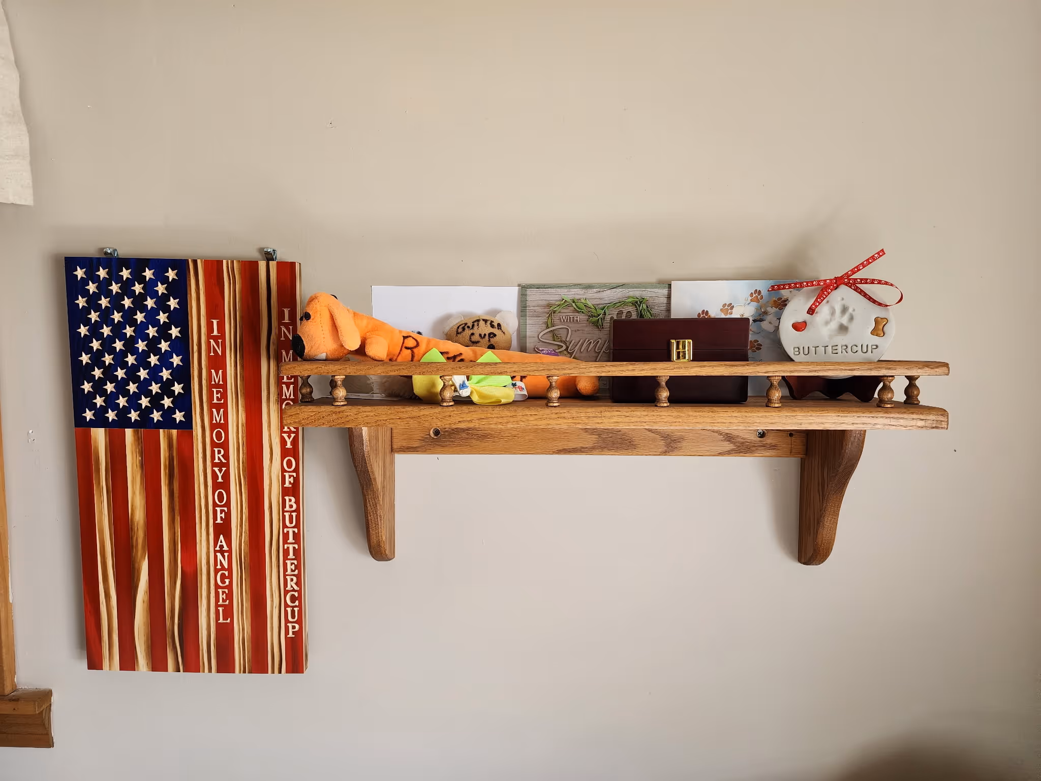 Wooden shelf with stuffed toy, urn, memorial plaques, and a flag reading in memory of Angel and Buttercup.