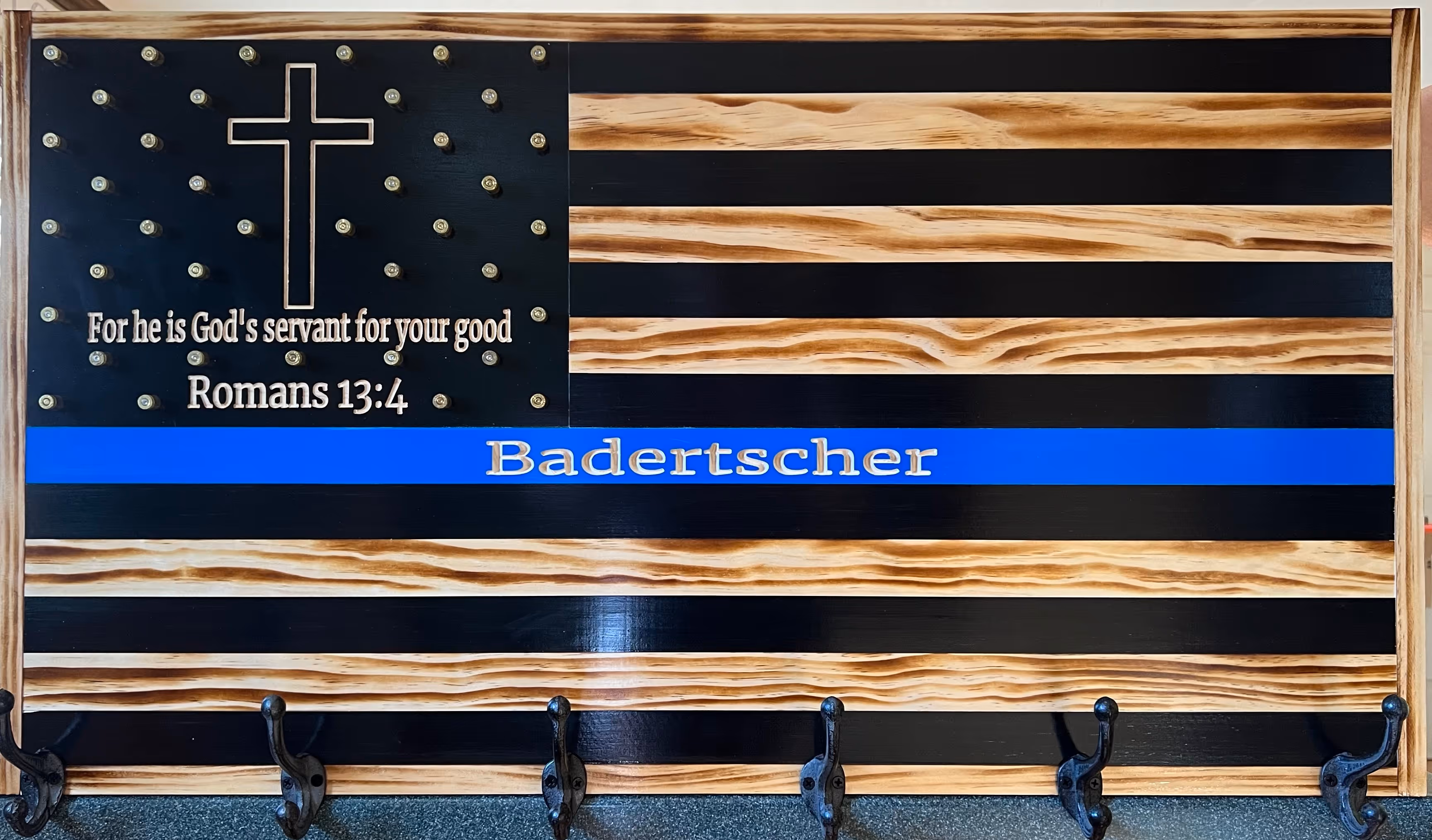 Wooden American flag with a cross, Bible verse Romans 13:4, a blue stripe, name Badertscher, and six hooks below.