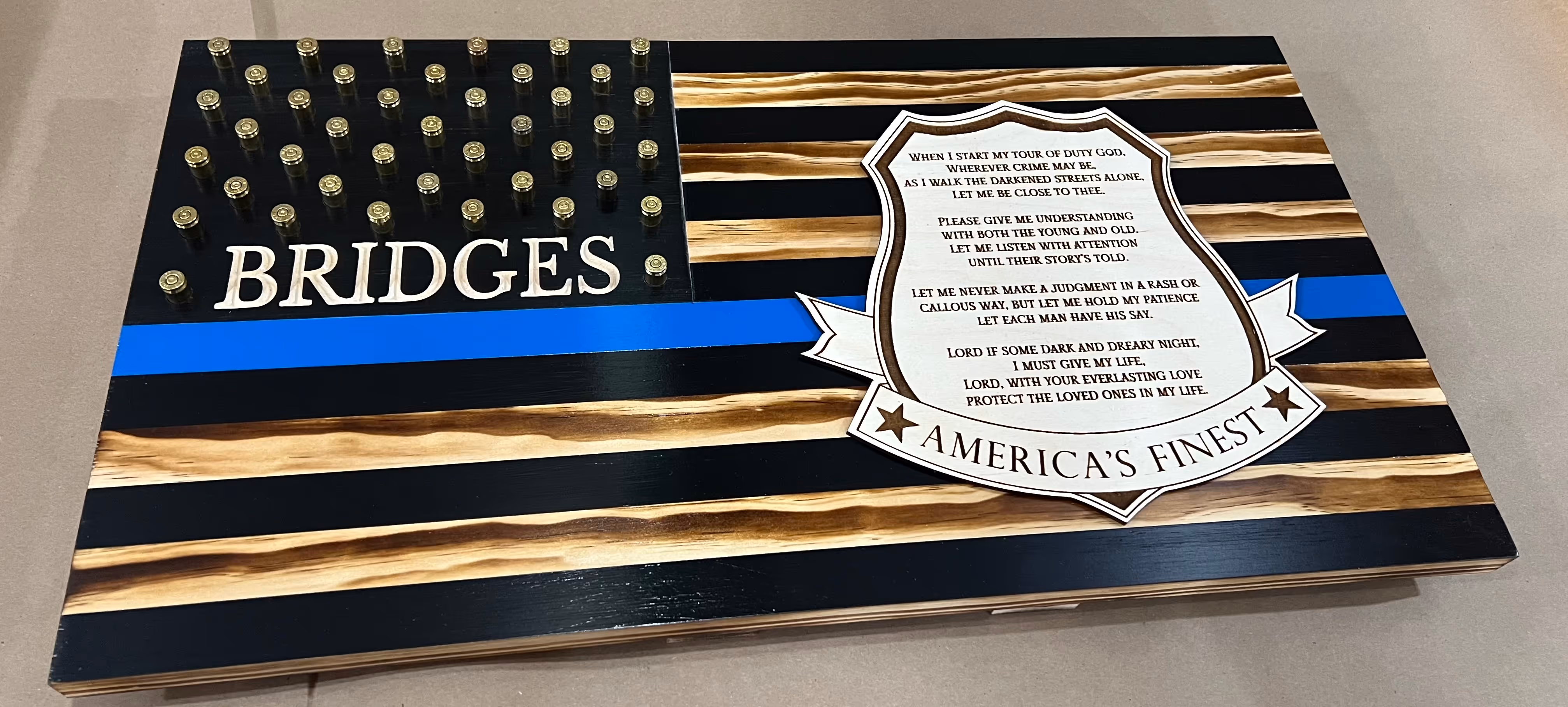 Wooden black and natural flag with blue stripe, bullet casings, 'BRIDGES,' and a shield with a police prayer.