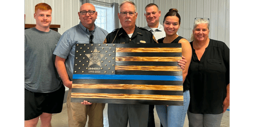 Five people stand together holding a burnt wood Thin Blue Line flag honoring Kimberly, 1993-2026.
