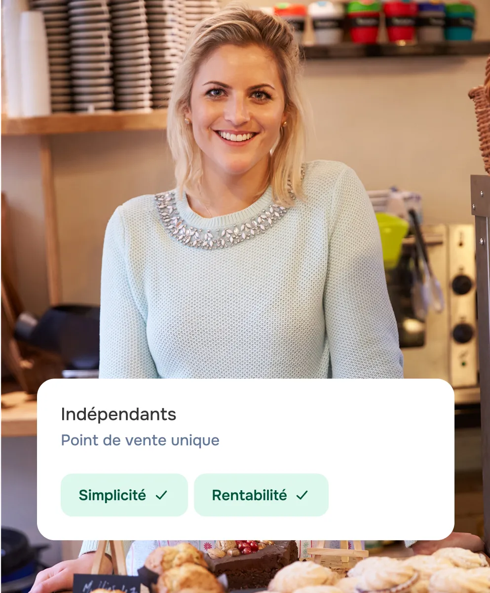 Femme blonde souriante portant un pull orné de bijoux, debout derrière un comptoir avec des pâtisseries.