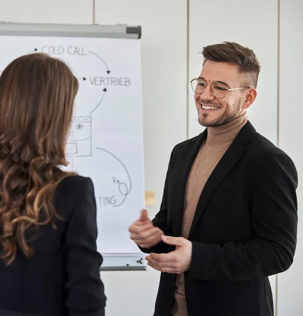 Zwei Personen führen ein Gespräch vor einem Flipchart mit Verkaufskonzepten wie Cold Call und Vertrieb.