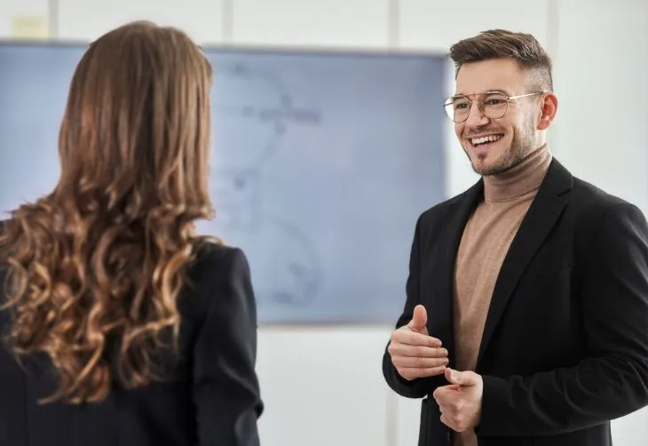 Ein Mann mit Brille und braunem Rollkragenpullover spricht lächelnd mit einer Frau vor einem Whiteboard.