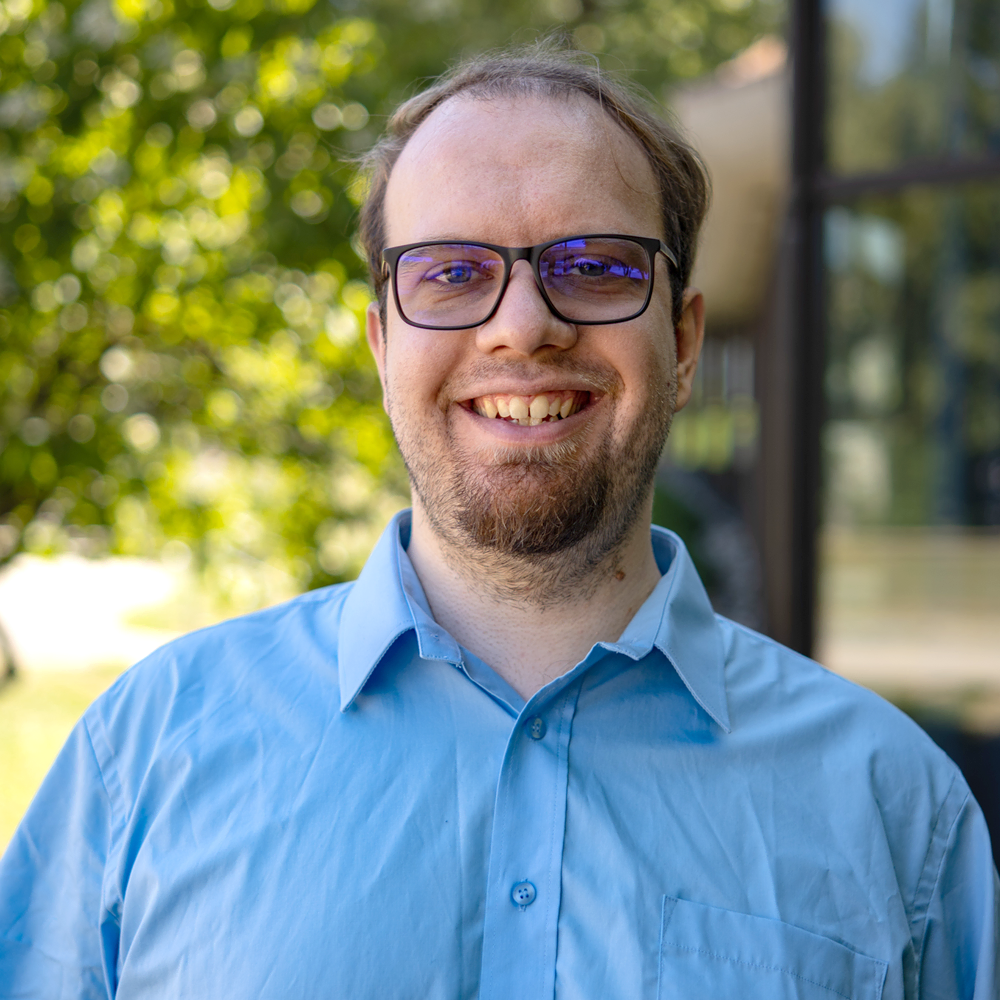 Porträt eines lächelnden Mannes mit Brille und blauem Hemd vor einem Hintergrund aus grünen Bäumen und Gebäudefenstern.