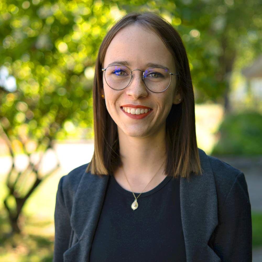 Junge Frau mit Brille, kurzen braunen Haaren und goldener Halskette, lächelnd vor grünem, unscharfem Hintergrund.