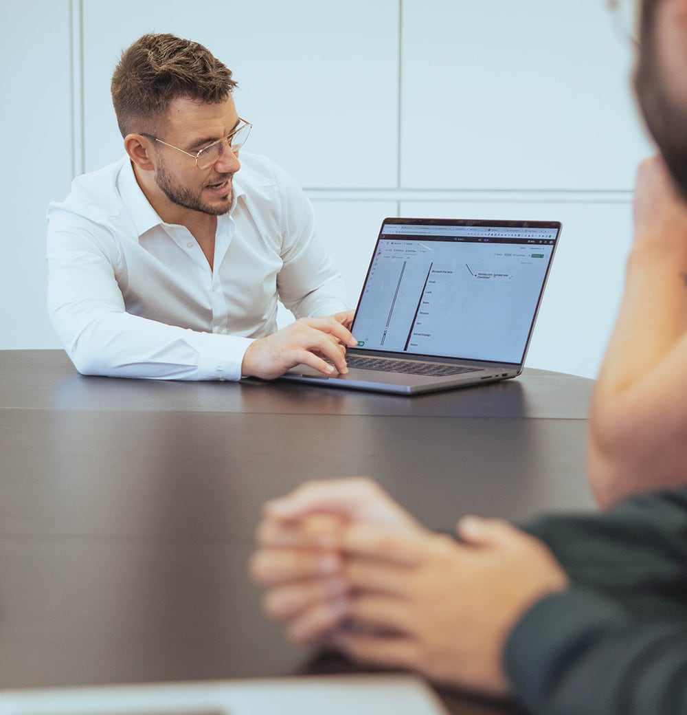 Ein Mann in weißem Hemd präsentiert in einem Meeting eine Analyse auf seinem Laptop mit Diagrammen und Daten.
