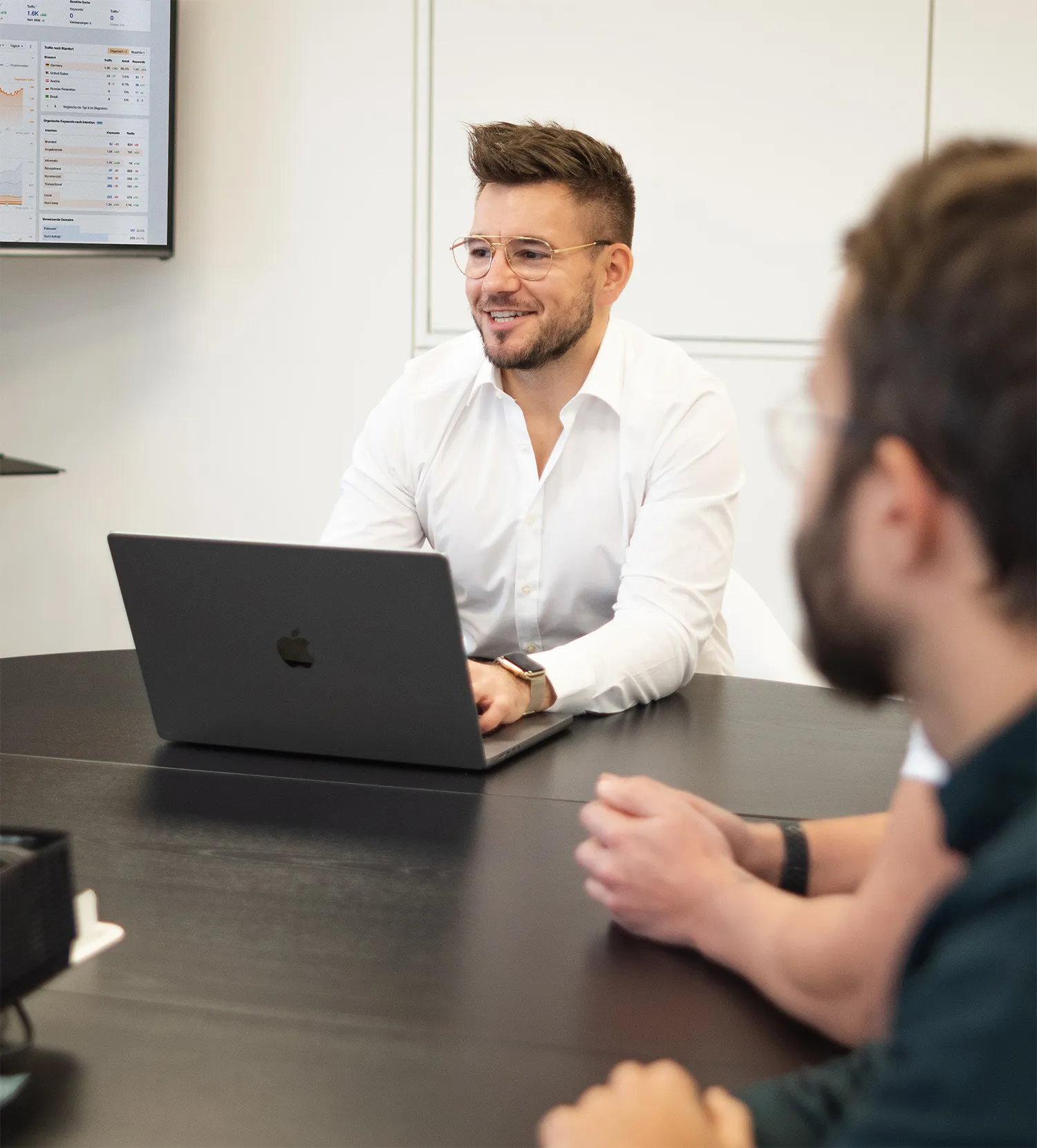 Mann mit weißem Hemd und Brille sitzt an einem Konferenztisch und lächelt vor einem Laptop, während zwei weitere Personen im Vordergrund zuhören.