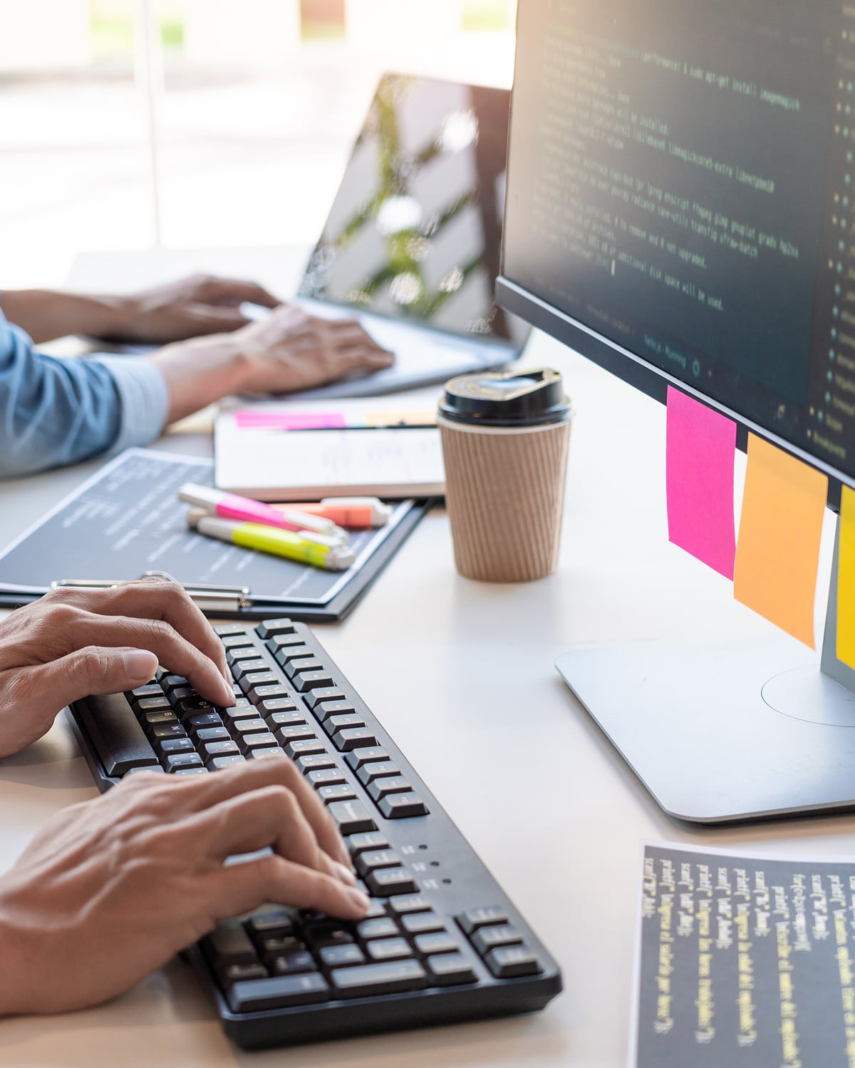 Person tippt an einer schwarzen Tastatur, im Hintergrund arbeitet eine weitere Person an einem Laptop, Monitor mit Code und bunte Haftnotizen sichtbar.