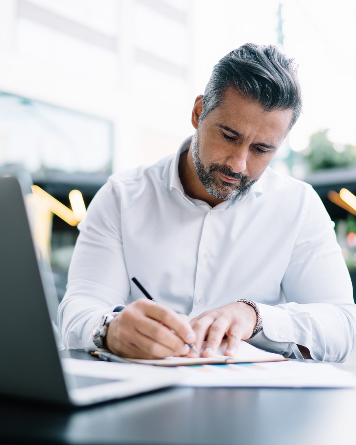 Mann mit grauem Haar und weißem Hemd schreibt konzentriert auf einem Tisch neben einem Laptop.