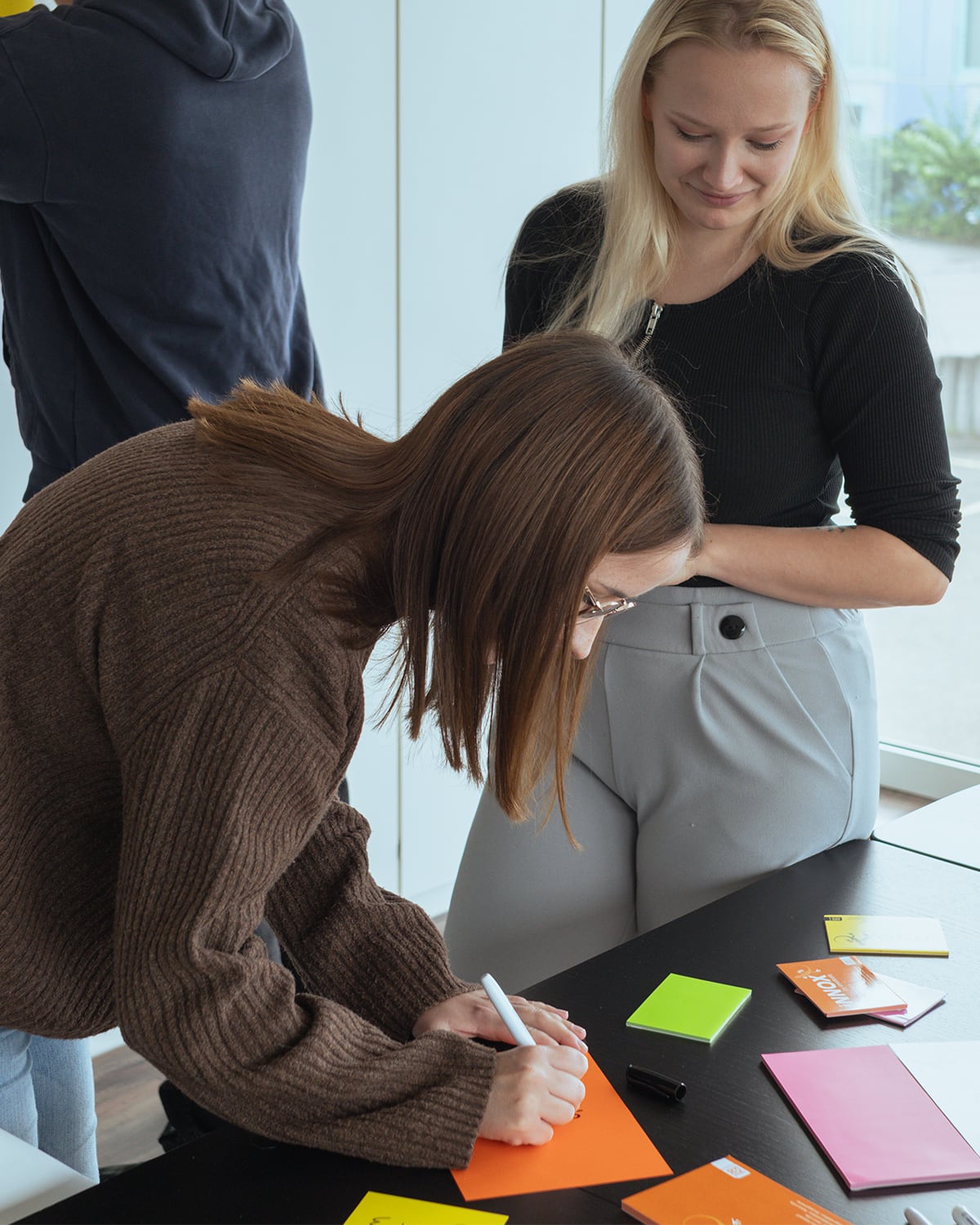Eine Frau mit braunem Pullover schreibt auf ein orangefarbenes Blatt, während eine blonde Frau neben ihr lächelnd zuschaut.