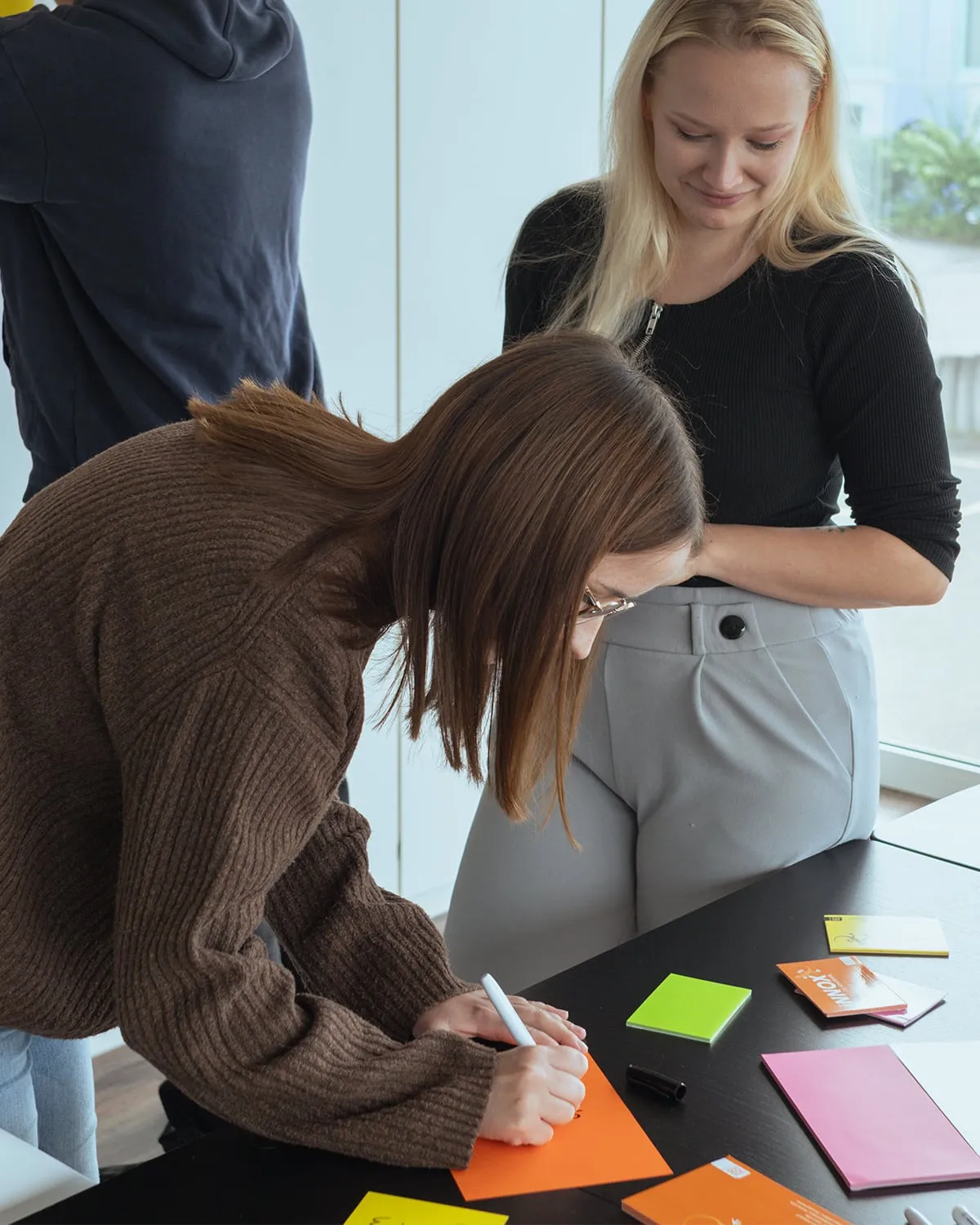 Eine Frau mit braunem Pullover schreibt auf ein orangefarbenes Blatt, während eine blonde Frau neben ihr lächelnd zuschaut.