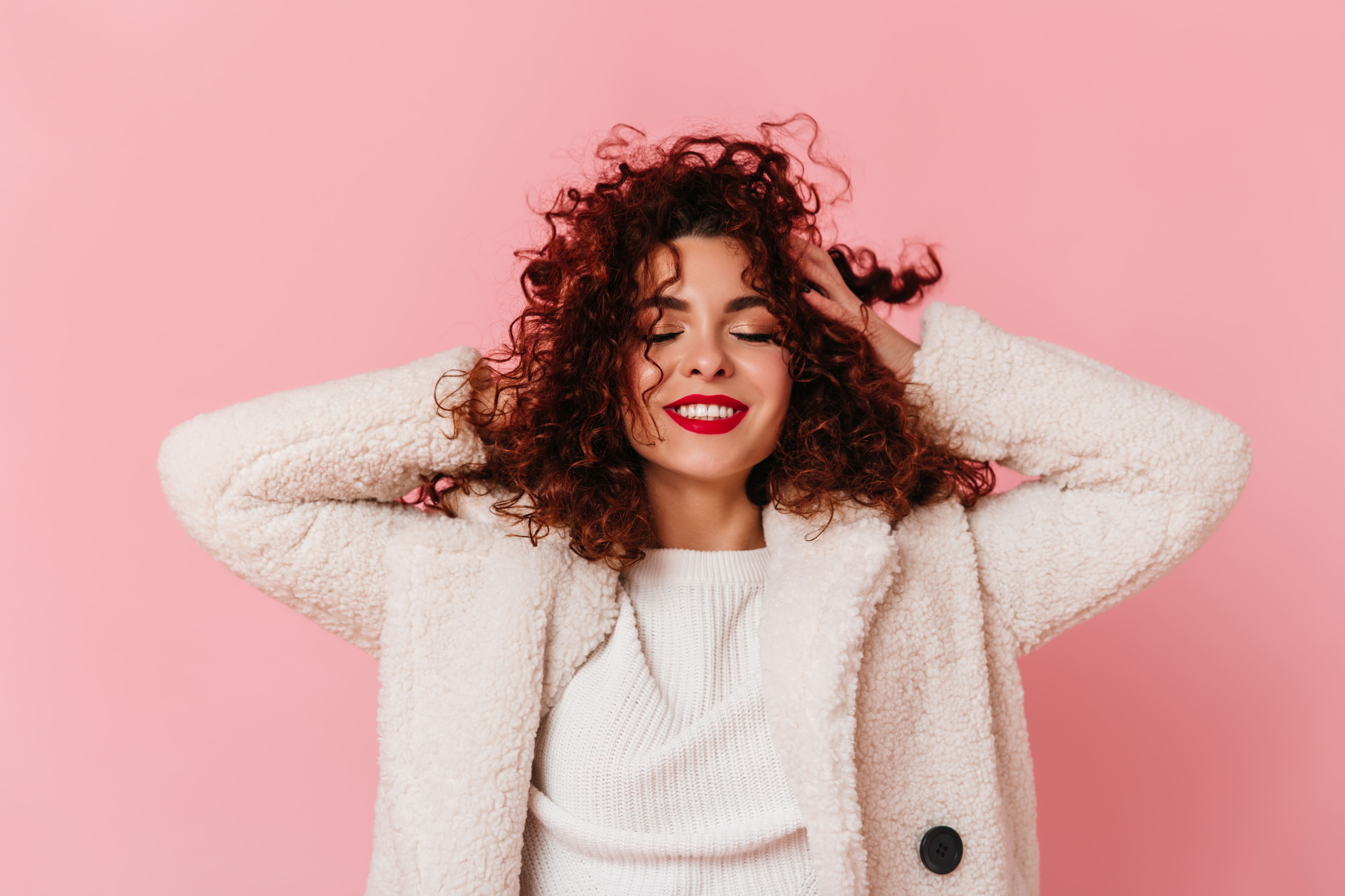 Woman with curly hair smiling against a pink background, illustrating modern beauty and haircare trends.
