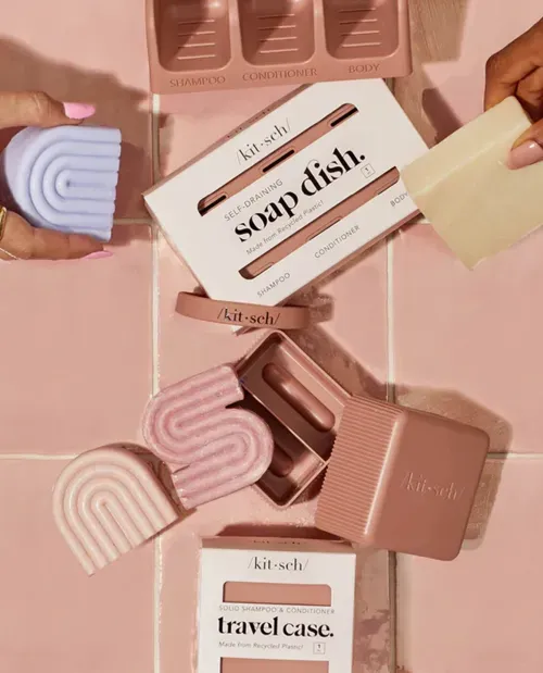 Flat lay of pink and lavender Kitsch brand solid shampoo and conditioner bars, a self-draining soap dish, and a travel case on pink tiled surface.