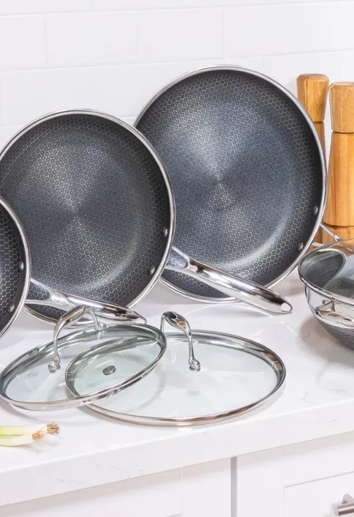 Three nonstick frying pans with hexagonal patterned interiors, two glass lids, and wooden salt and pepper grinders on a white kitchen countertop.