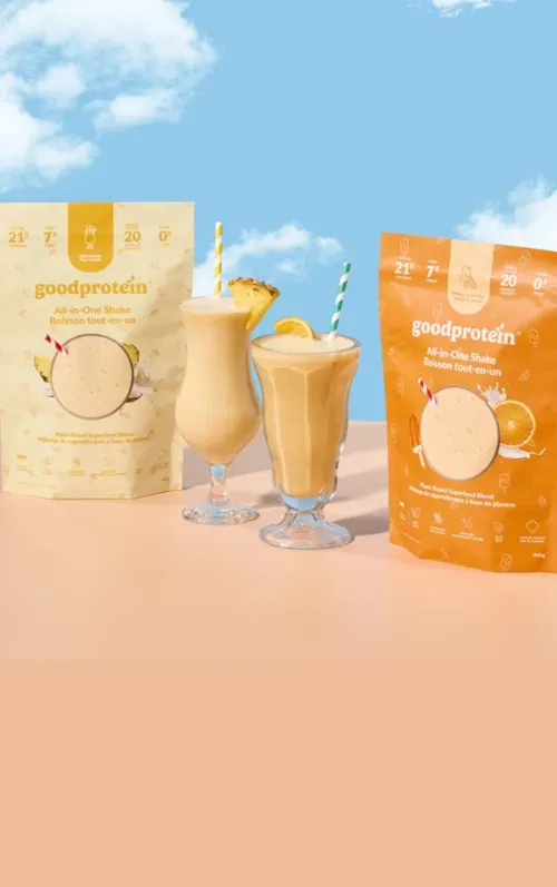 Two bags of goodprotein All-in-One Shake plant-based superfood blend with two creamy shakes garnished with pineapple and orange slices and striped straws.