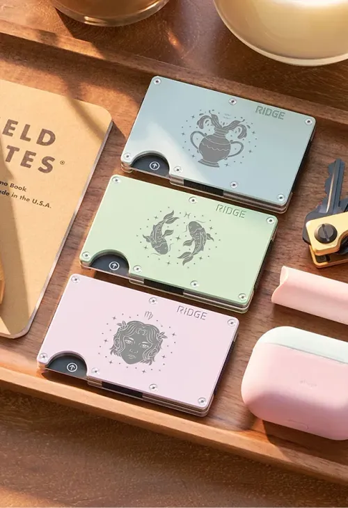 Three Ridge brand slim wallets in pastel colors with zodiac illustrations of Aquarius, Pisces, and Virgo on a wooden tray.