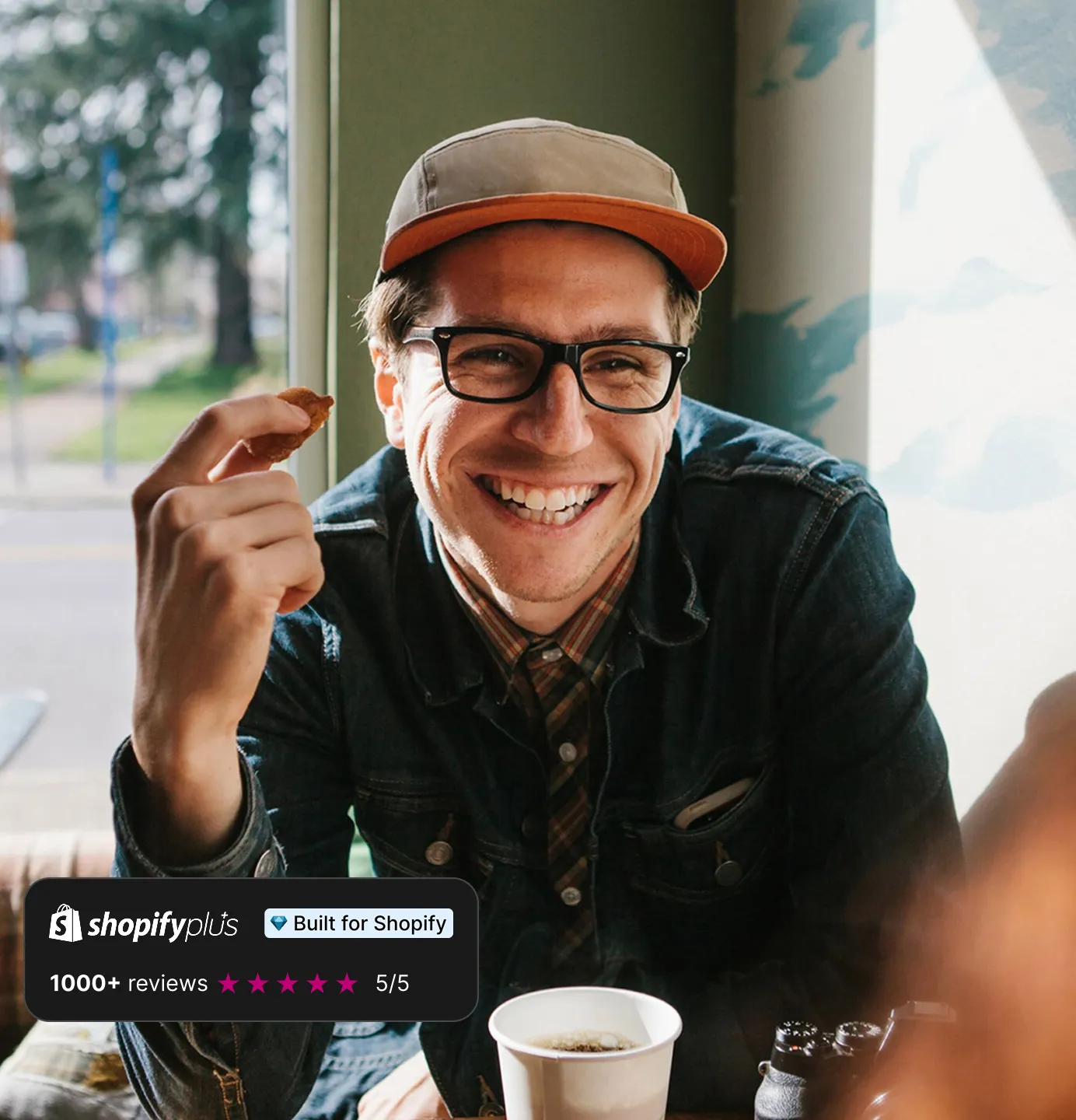 Smiling man wearing glasses and a cap eating a snack with a cup of coffee in front of him indoors.