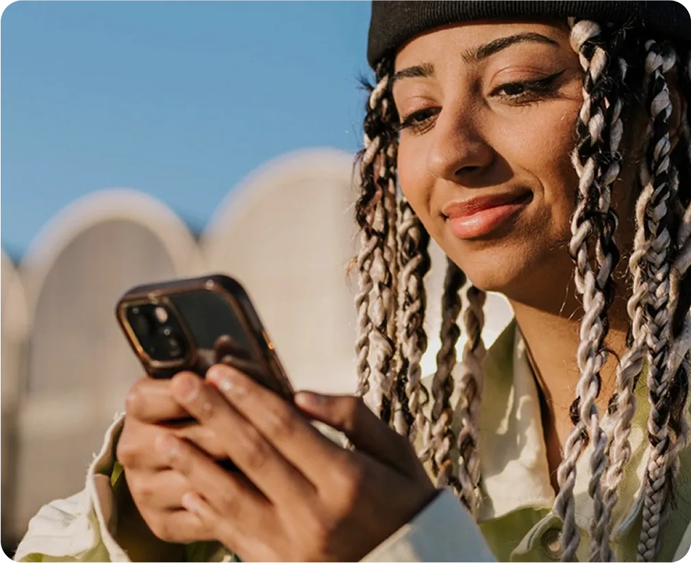 Smiling woman with braided hair and a beanie looking at her smartphone outdoors against a clear blue sky.