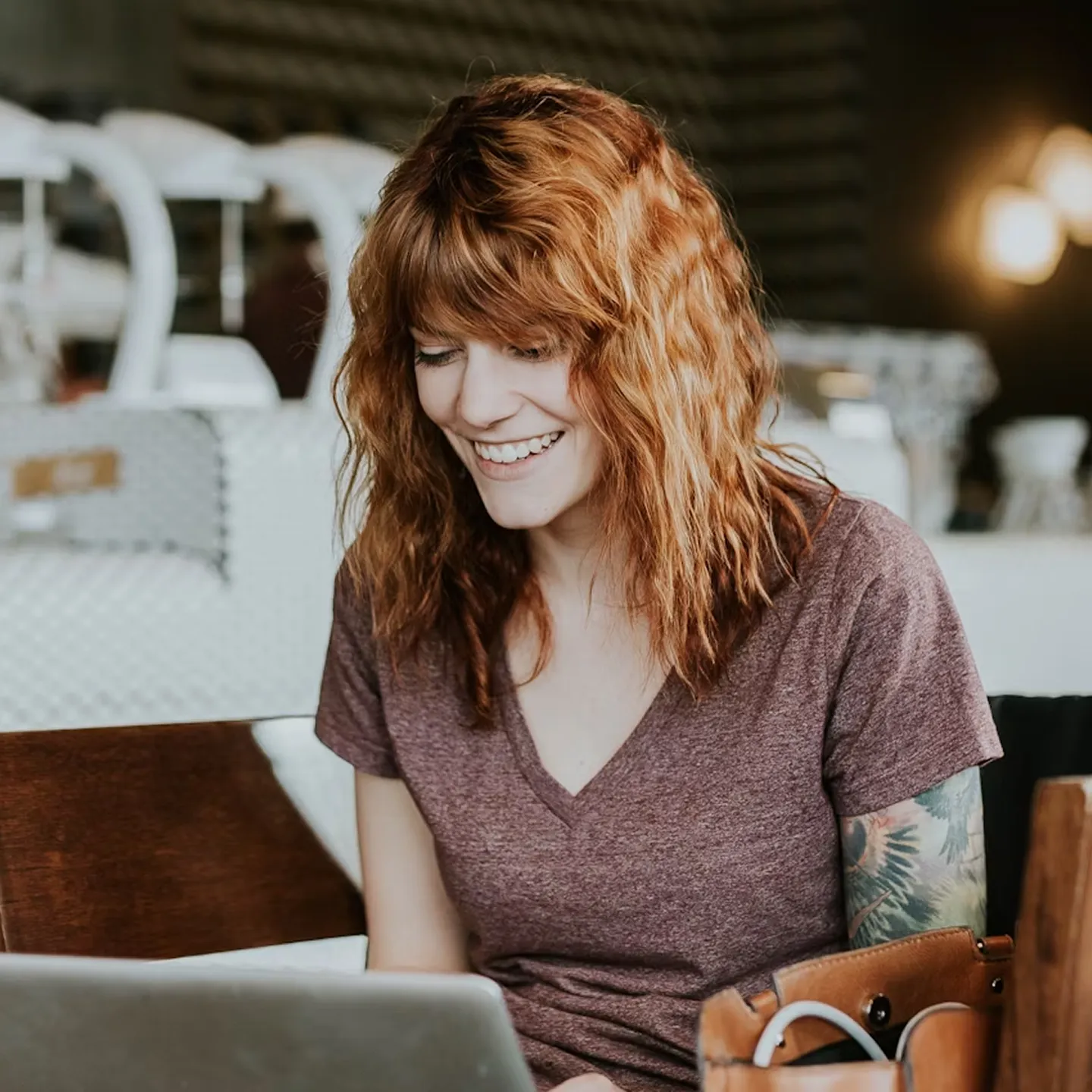 Smiling woman with red hair and a tattoo on her arm working on a laptop in a cozy café.