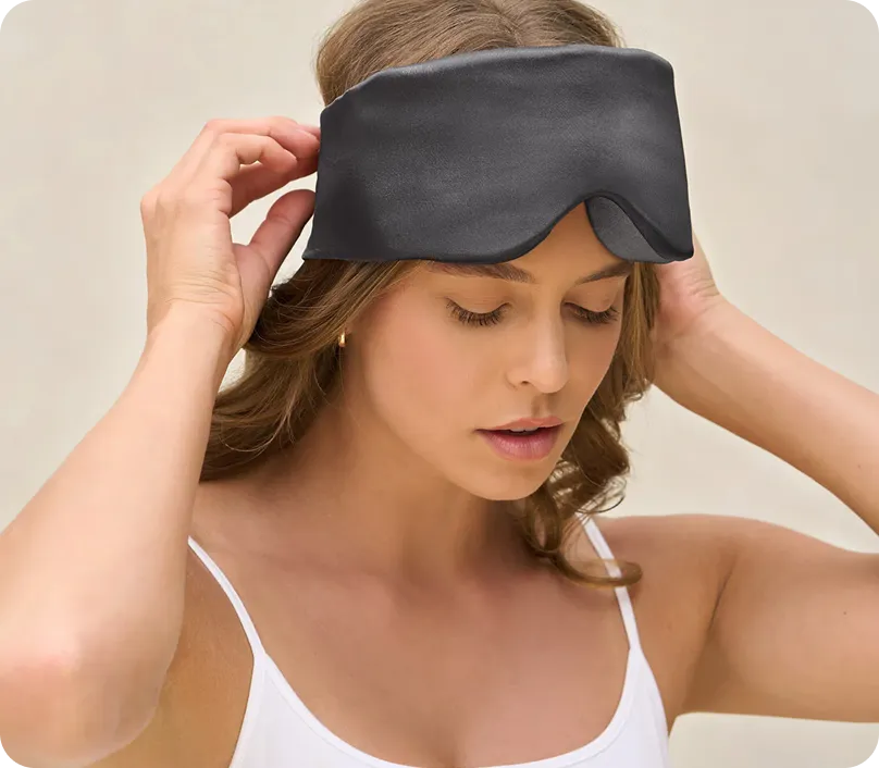 Woman adjusting a black sleep mask on her forehead wearing a white tank top.
