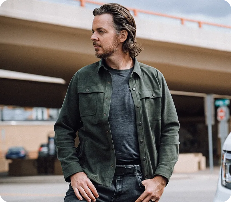 Man with medium-length hair and a beard wearing a green jacket over a dark gray shirt standing outdoors near a road and overpass.