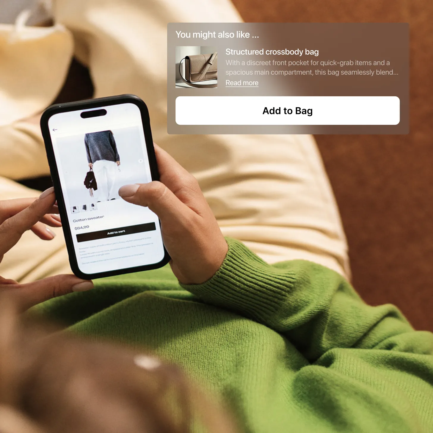 Person in green sweater browsing an online store on a smartphone, viewing a cotton sweater priced at $84.99 with options to add to cart and a suggestion for a structured crossbody bag.