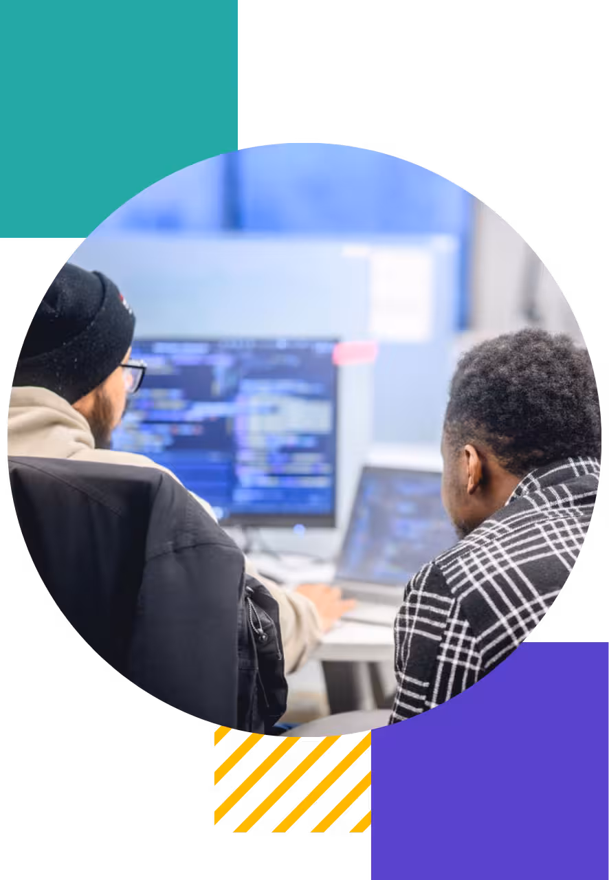Two developers reviewing code on a desktop monitor and laptop in a coworking space, with Genesis brand shapes in blue, yellow, and red in the background.