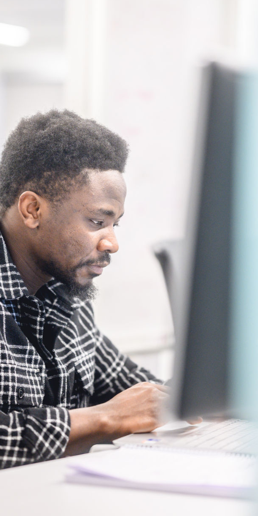 Man with a beard working at a desktop computer, plaid flannel shirt, notebook open beside the keyboard.