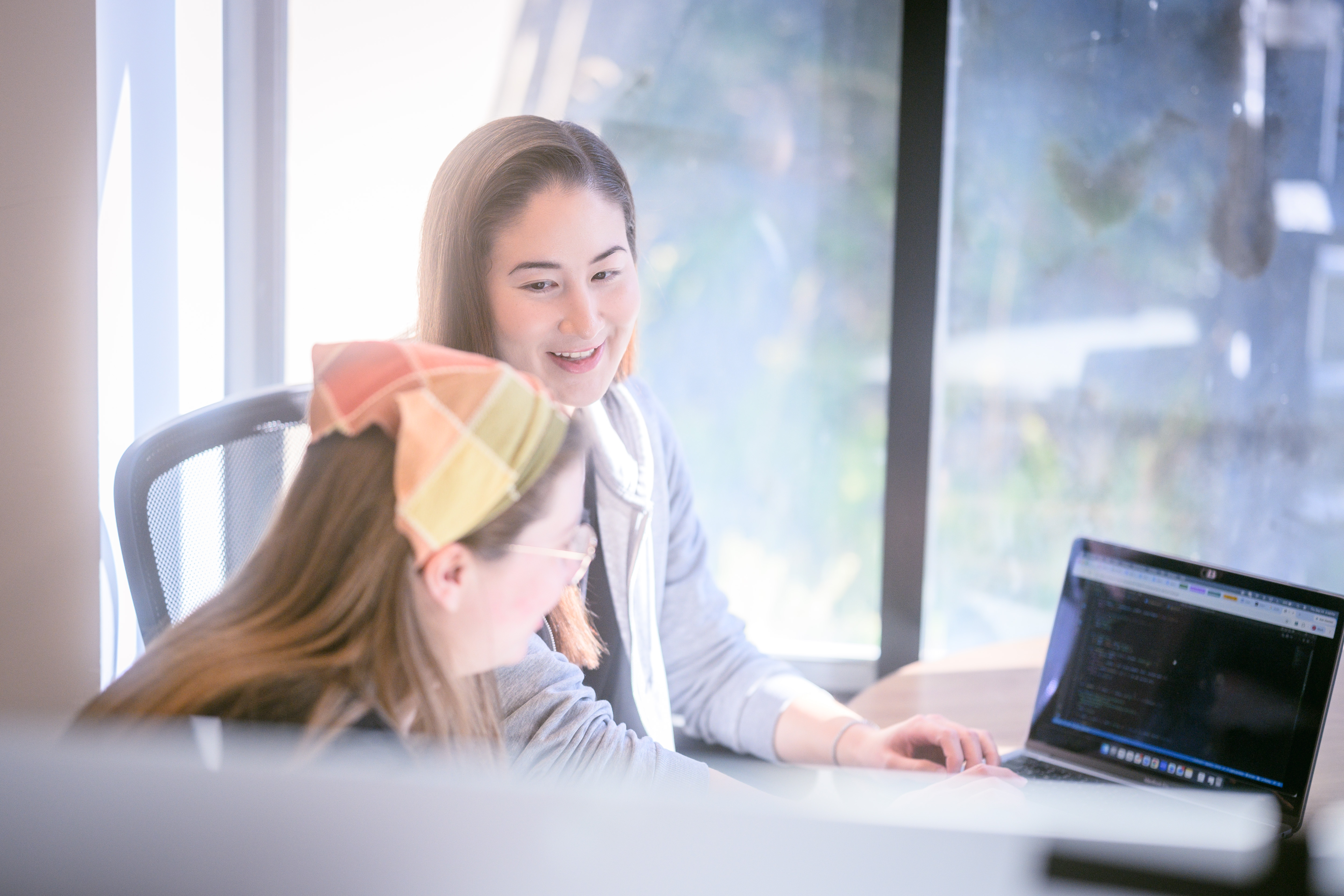 Two women smiling at a laptop showing code, sitting near a bright window, one wearing a plaid cap.