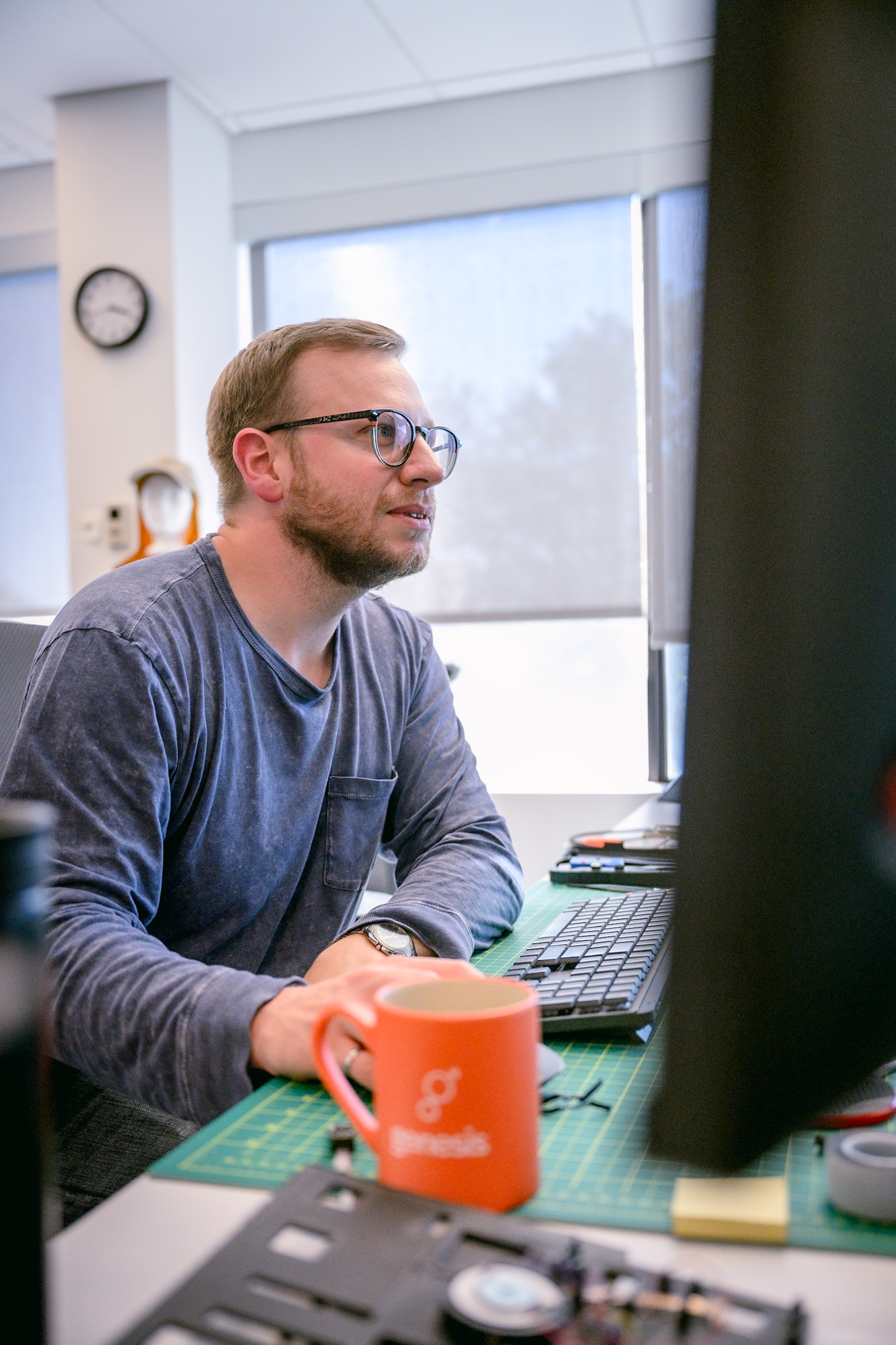 Man with glasses seated at a desk with dual monitors, a Genesis-branded mug, and electronics on a green cutting mat.