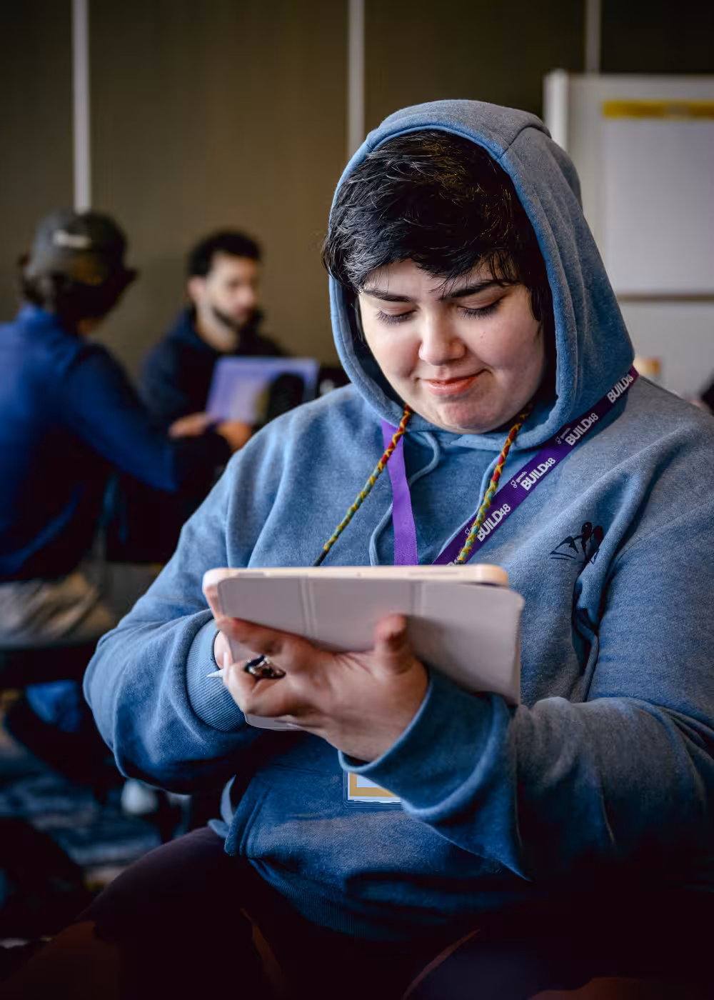 Woman with glasses smiling while typing on a laptop, sitting at a bright desk near a window.