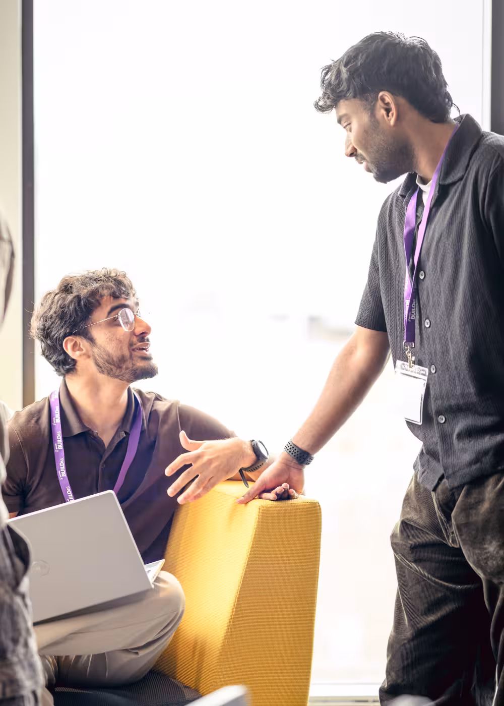 Two men wearing BUILD48 lanyards talking near a bright window, one seated with a laptop and gesturing, the other standing and listening.