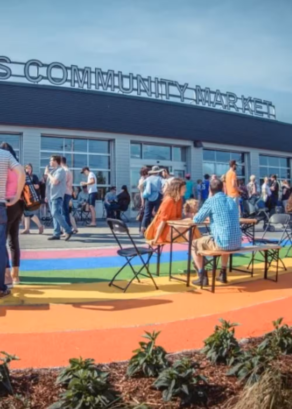 Crowd of people outside the Community Market building, sitting and standing near picnic tables on a rainbow-painted walkway.