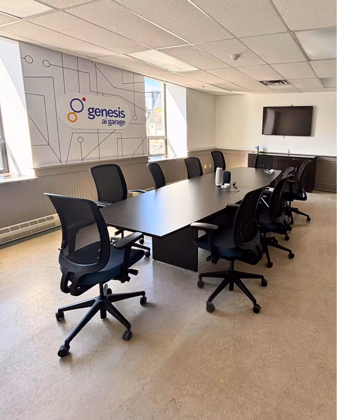 Boardroom at Genesis AI Garage with a long dark conference table, ten black mesh office chairs, a wall-mounted TV, and the Genesis AI Garage logo on a circuit-pattern wall.