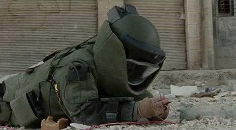 Bomb squad member wearing a protective suit and helmet lying on rubble while handling a device.
