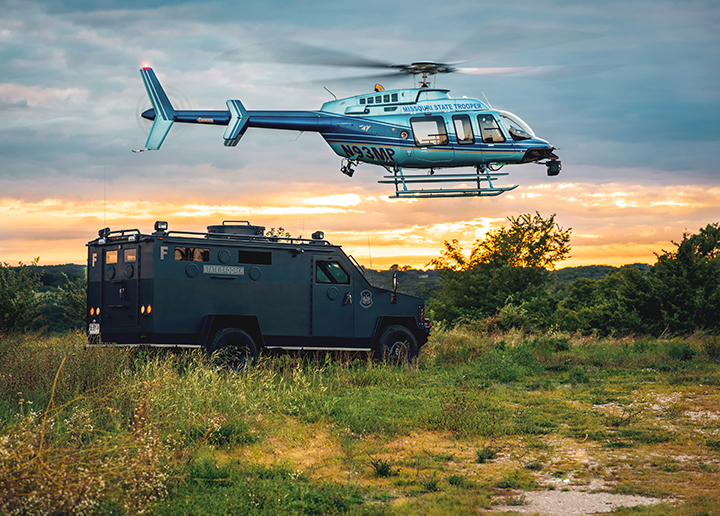 Missouri State Highway Patrol helicopter flying above an armored vehicle in an open grassy area at sunset.