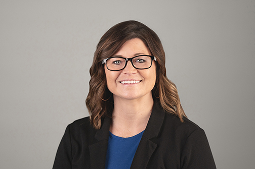 Smiling woman with brown hair wearing glasses, a black blazer, and a blue top, against a plain gray background.