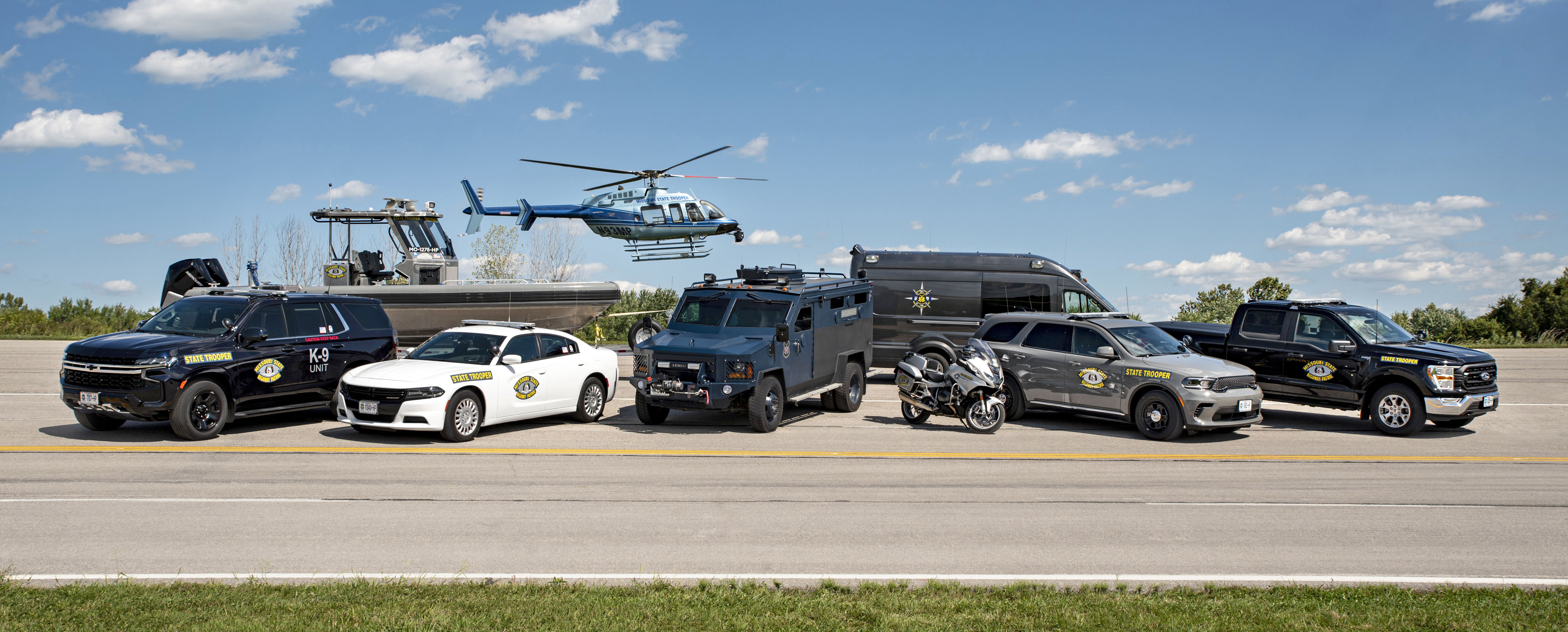 Five parked law enforcement vehicles in a row on a concrete surface, including SUVs, sedans, and a pickup truck with official badges.