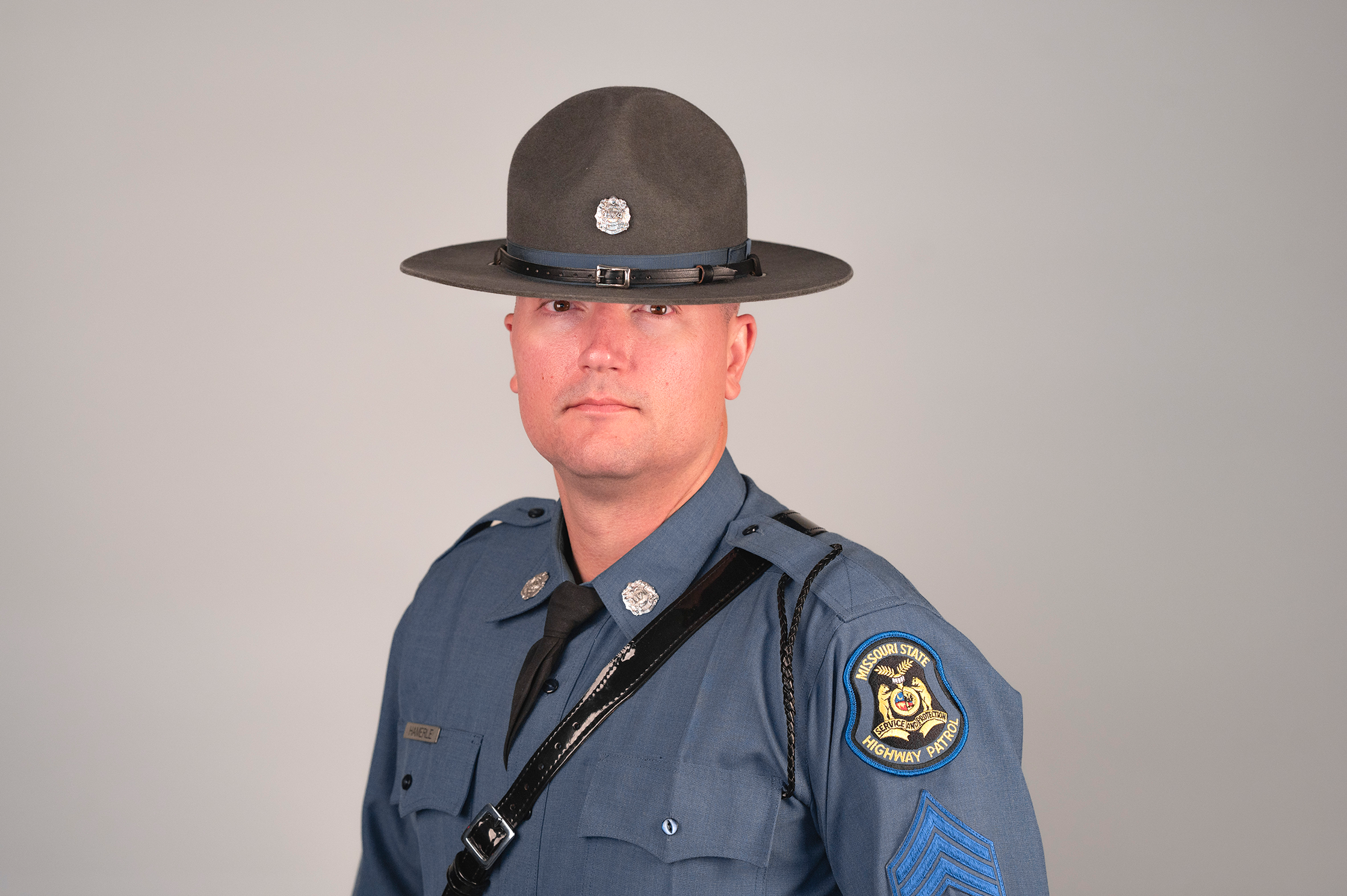 Highway Patrol officer in a blue-gray uniform shirt and black tie, wearing a gray campaign hat with a badge, against a plain gray background.