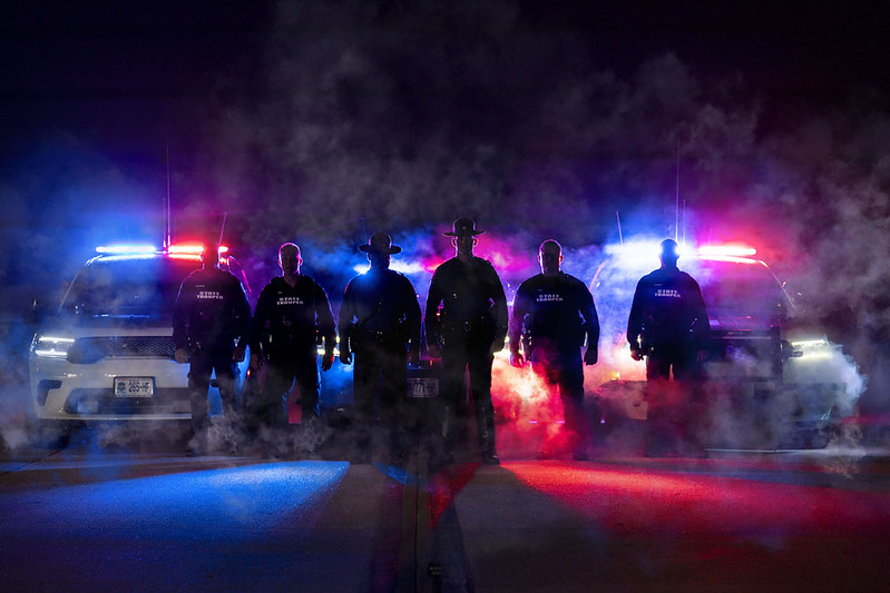 Six members of the Storm team stand in silhouette in front of patrol vehicles, lights and fog.