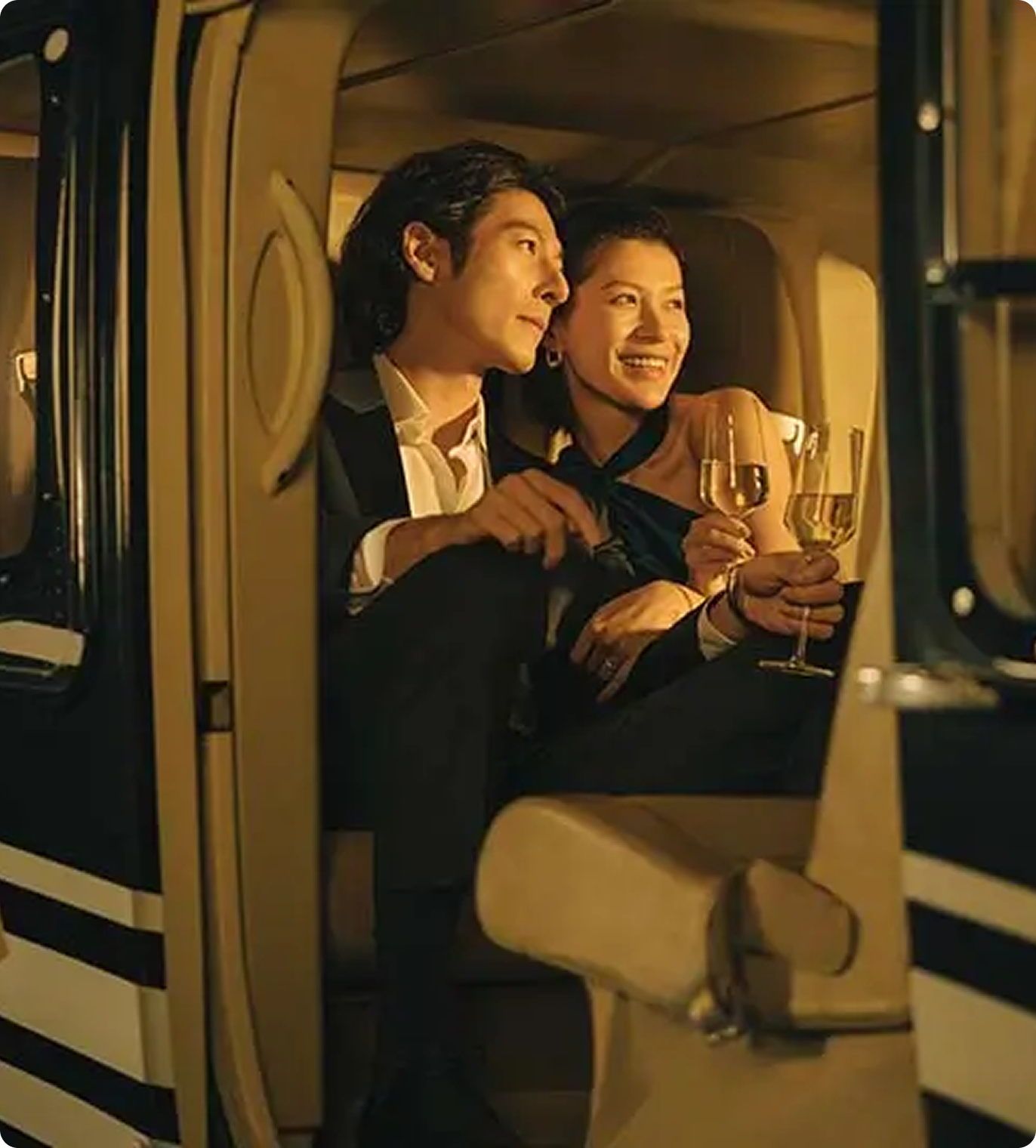Smiling couple dressed formally, sitting inside a luxury helicopter and holding glasses of white wine.