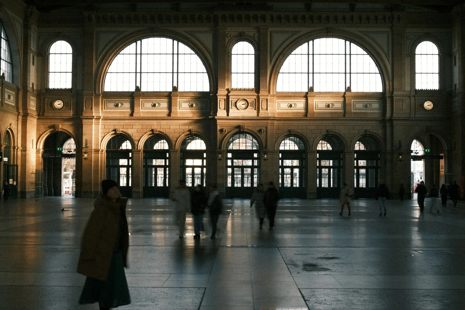 Hall de gare baigné de lumière du soir, silhouettes de voyageurs en mouvement