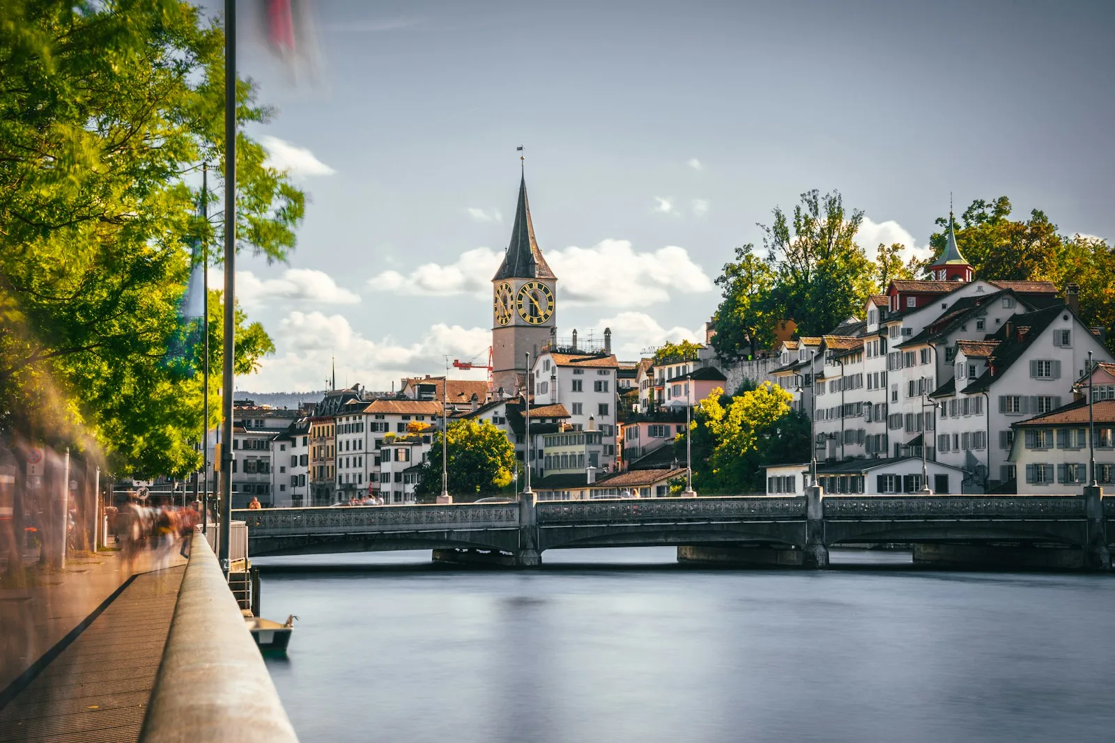 Quai de la Limmat à Zurich en fin de journée, le clocher de Saint-Pierre au fond et des passants en flou de mouvement sur le quai.