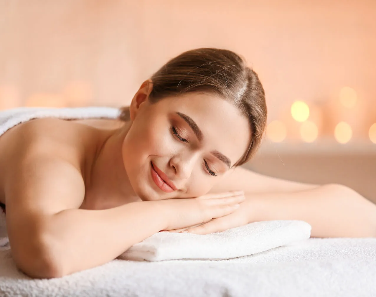 Smiling girl applying coffee mask or scrub on skin, looks at camera, wearing towel, doing beauty treatment manipulation, keeping finger on lips, posing against lilac wall.