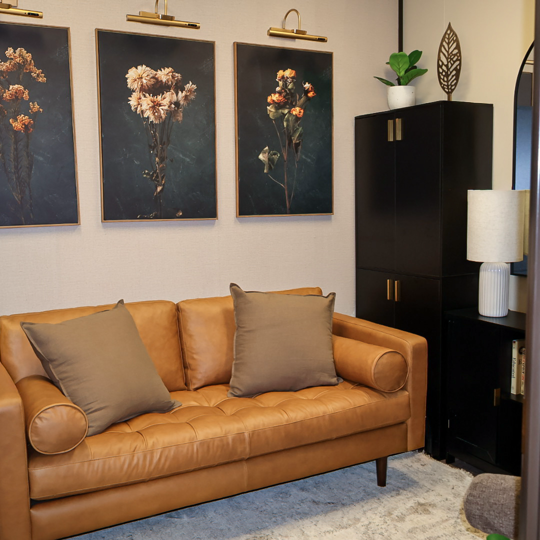 Cozy office corner with a tan leather couch, gray pillows, floral wall art, black cabinet, and table lamp.