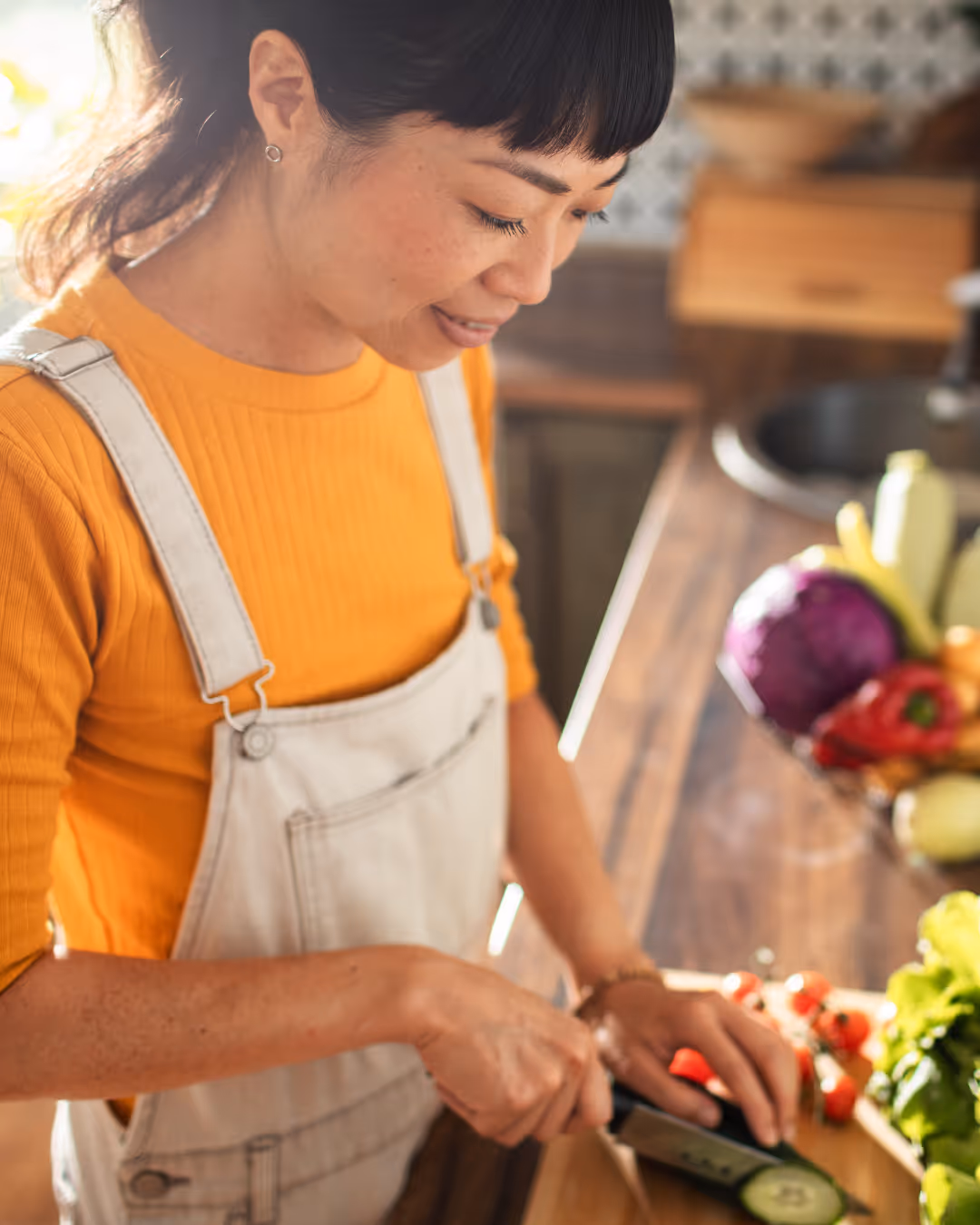 Woman in yellow shirt and overalls slicing a cucumber on a wooden cutting board in a kitchen.