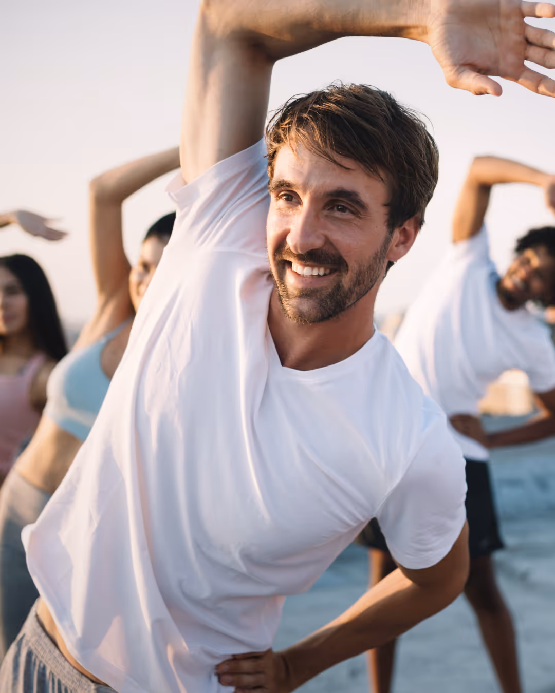 Smiling man in white t-shirt stretching with arms raised during outdoor group exercise.