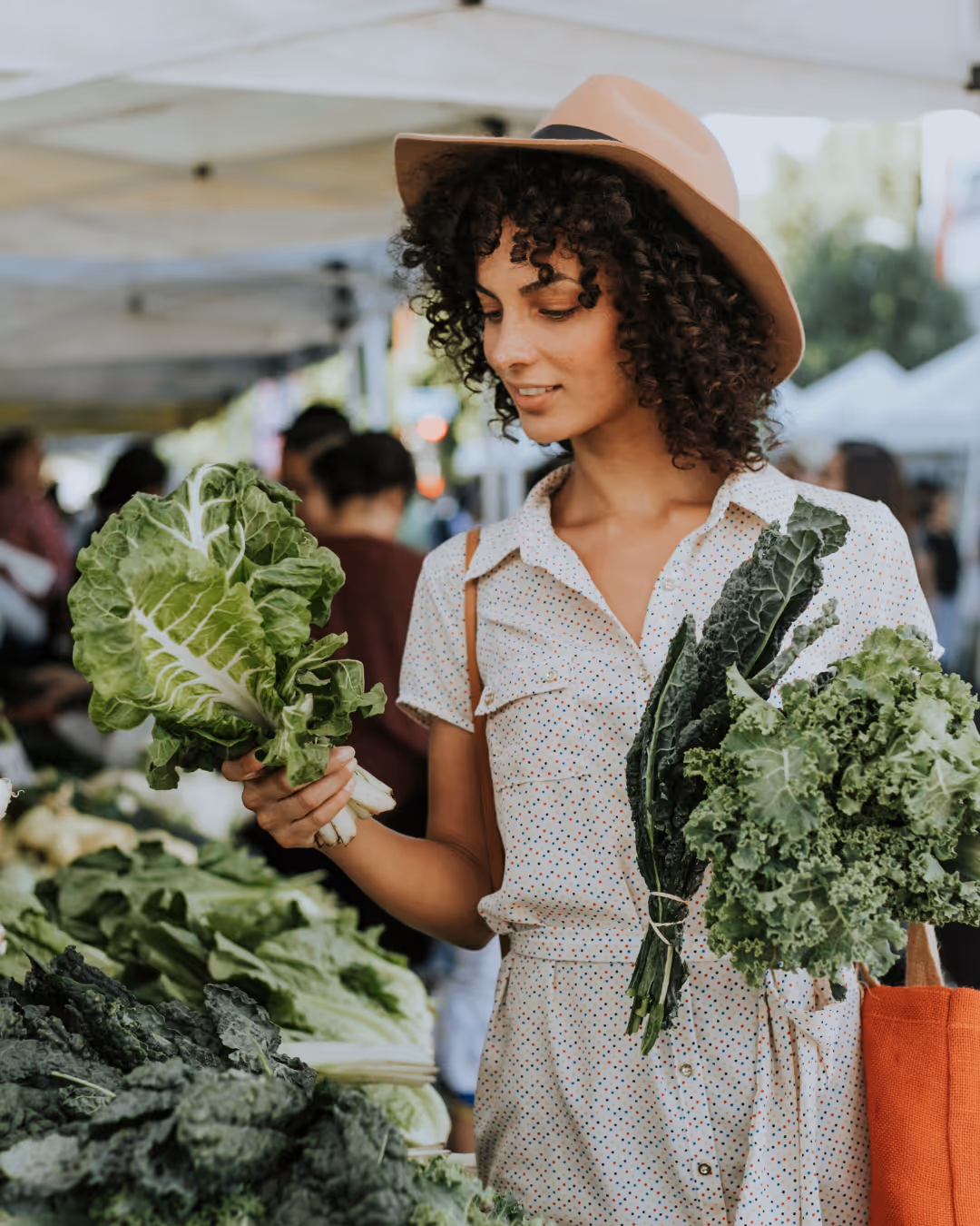Woman in a tan hat holding fresh leafy greens at a farmer's market stall.